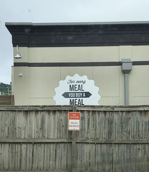 Marketing sticker from a restaurant that donates a meal to impoverished persons for every meal purchased in-store. Half of the sign is cut off by a fence, and what remains reads "FOR EVERY MEAL, YOU BUY A MEAL"